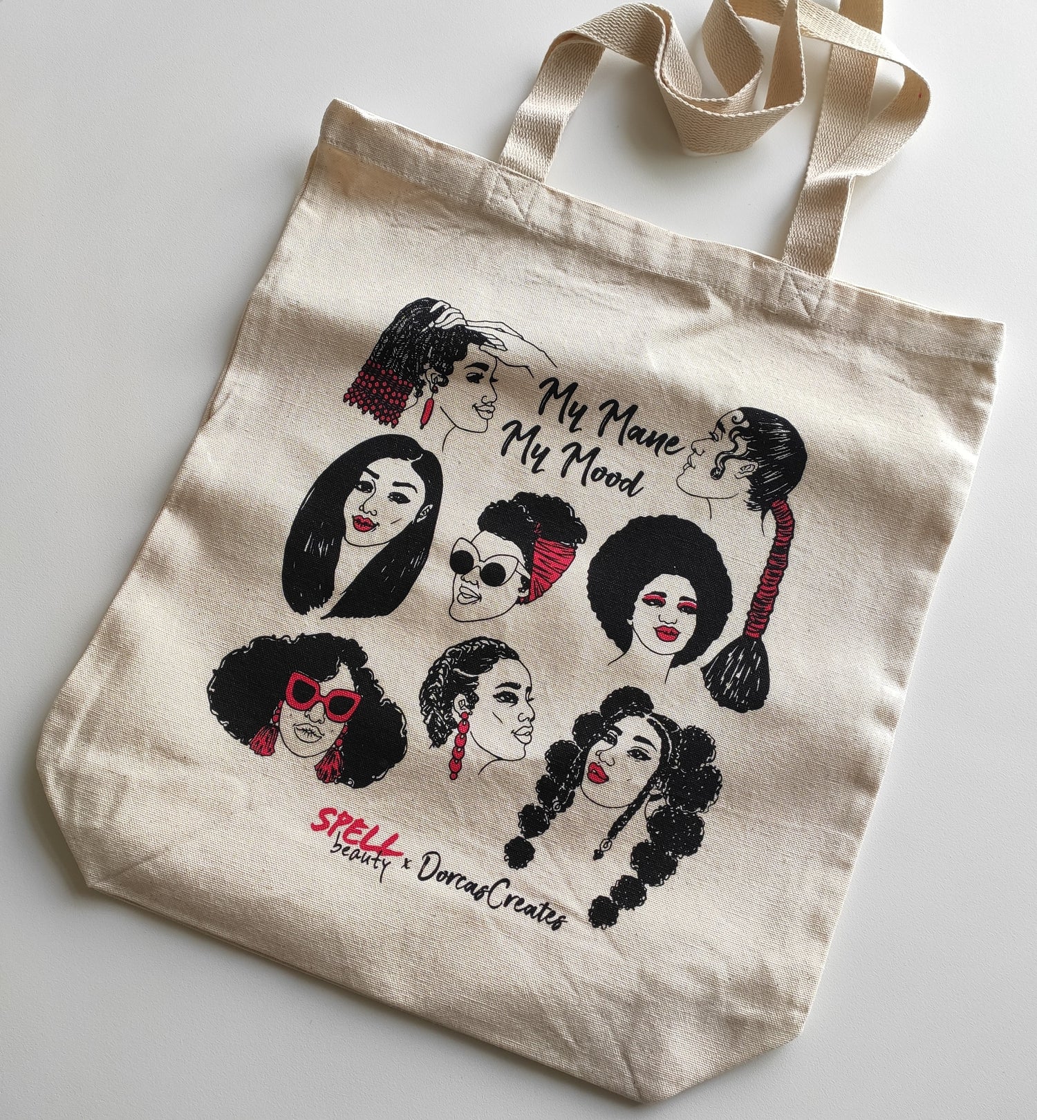 Natural coloured tote bag lying flat on white background. Tote bag features illustrations of 8 different hairstyles worn by Black women. Illustrations by Dorcas Magbadelo for Spell Beauty